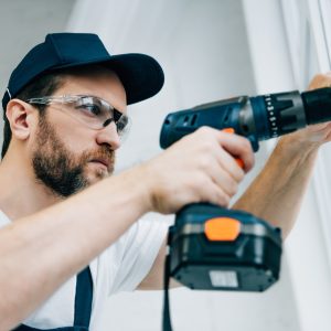 adult repairman in goggles fixing window handle by electric drill